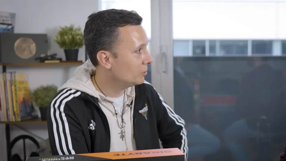 Daniel Pajuelo, con chaqueta negra y sudadera clara, sentado de perfil ante la mesa de montaje, detrás de la caja de la tarjeta gráfica.