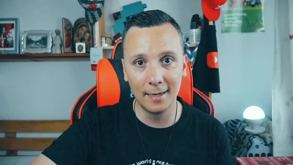 Daniel Pajuelo con camiseta negra hablando a cámara en su estudio, con estanterías y una lámpara detrás.